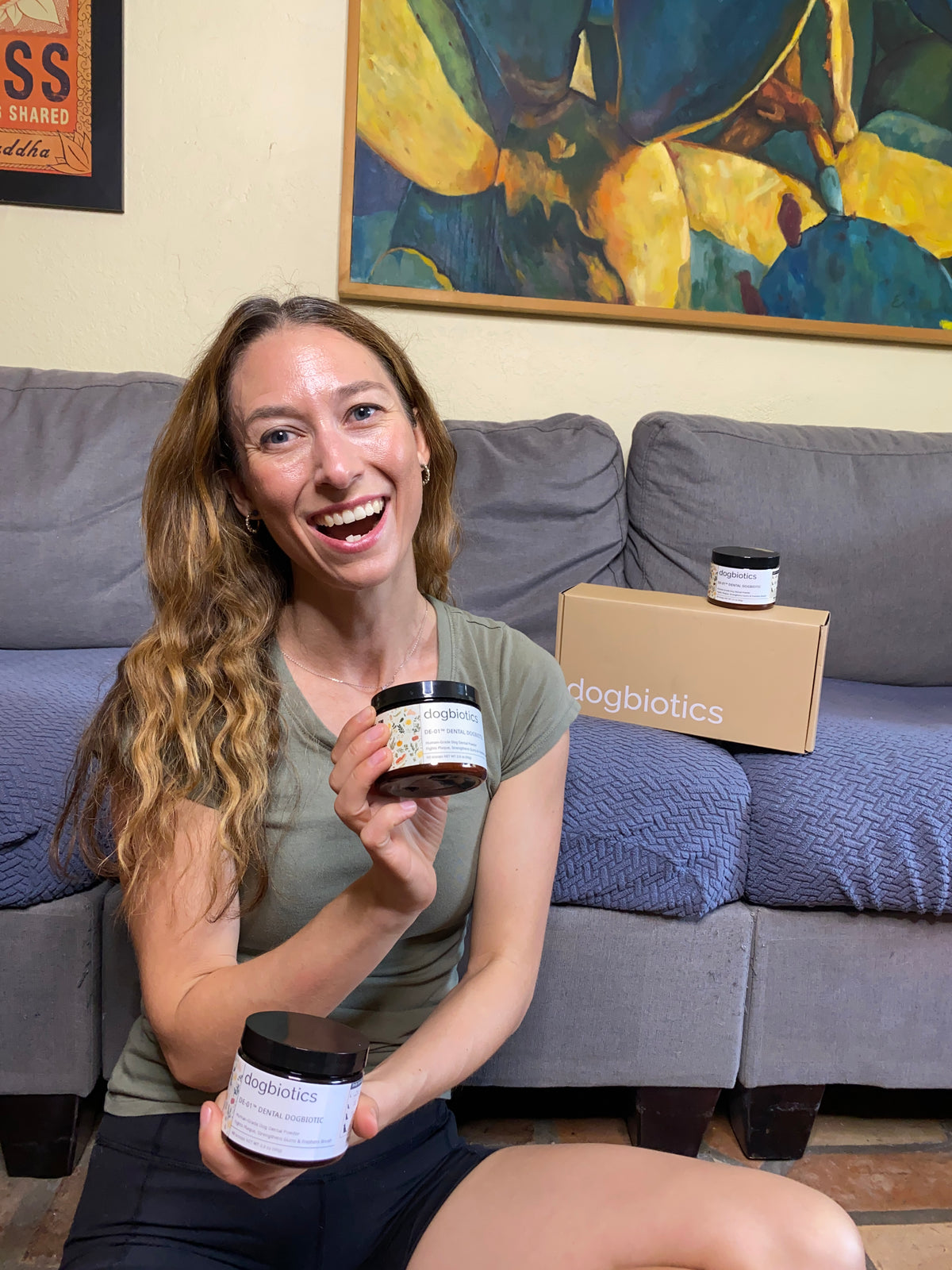 Smiling person holding products labeled 'dogbiotics' near a couch with colorful artwork behind.