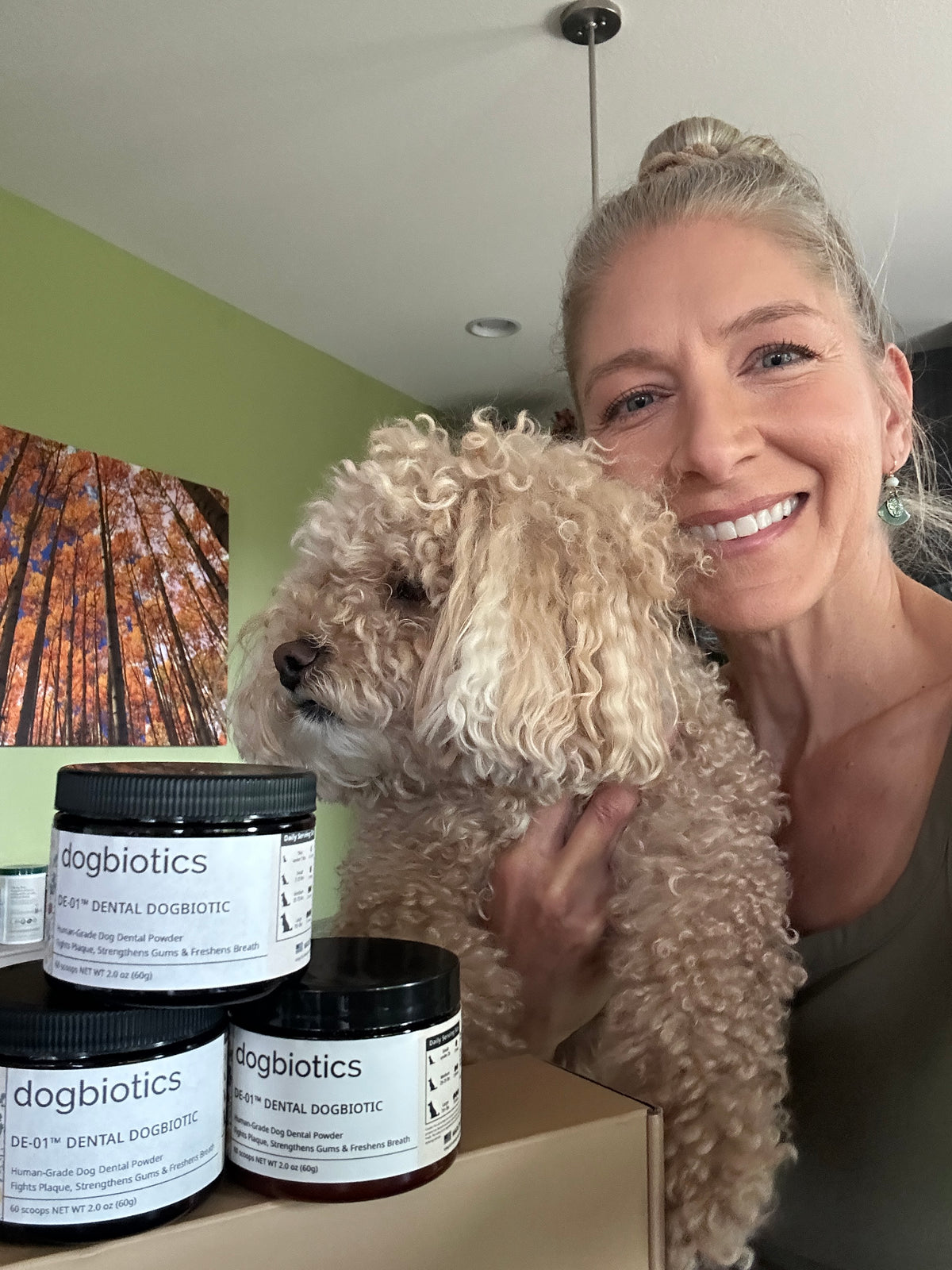 Smiling woman holds a fluffy dog beside jars labeled 'dogbiotics' in a room with green walls.